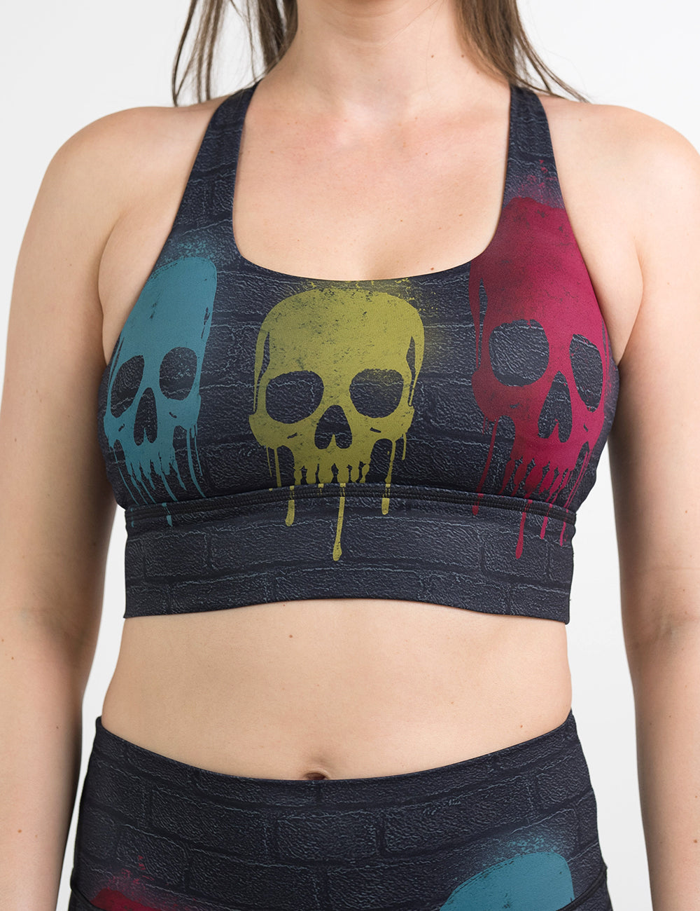 front of the black brick style longline bra with yellow, teal, and red skulls spray painted all over against a white background