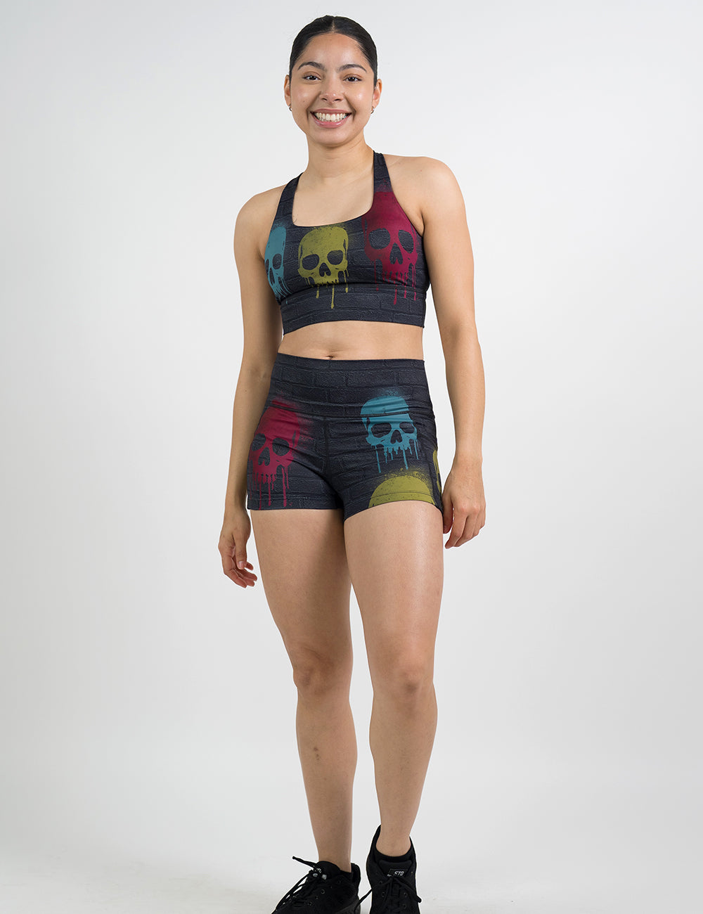 model wearing the 2.5 inch black brick pattern shorts and bra with spray painted yellow, blue and red skulls all over against a white background