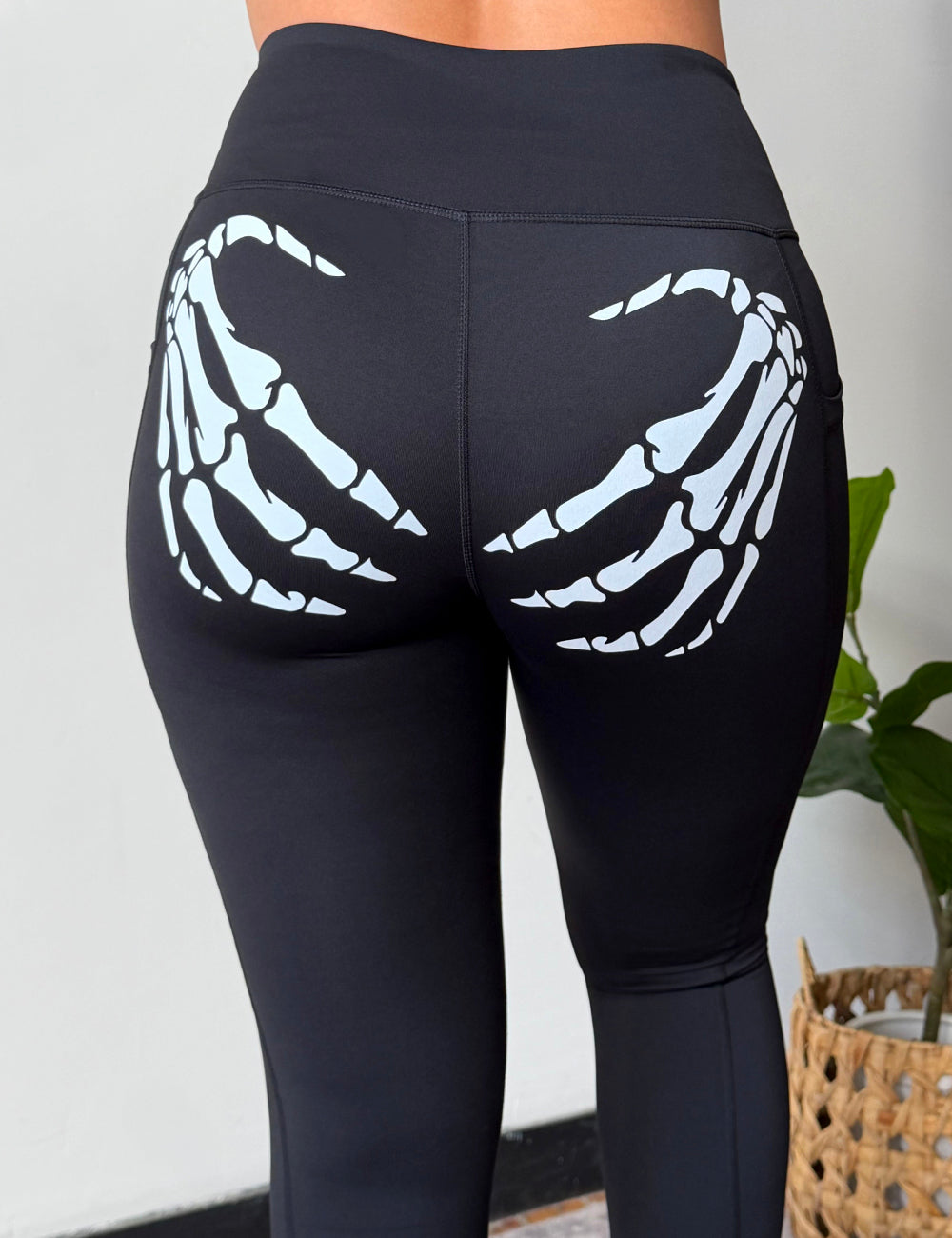 Black high-waisted leggings with a white skeleton hand graphic printed on the back of the thighs, shown on a model from behind in an indoor setting.