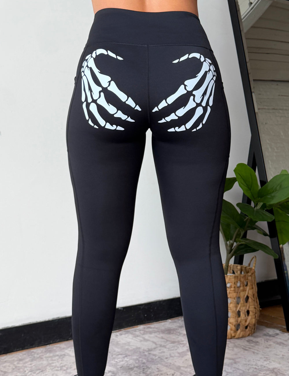 Black high-waisted leggings with a white skeleton hand graphic printed on the back of the thighs, shown on a model from behind in an indoor setting.