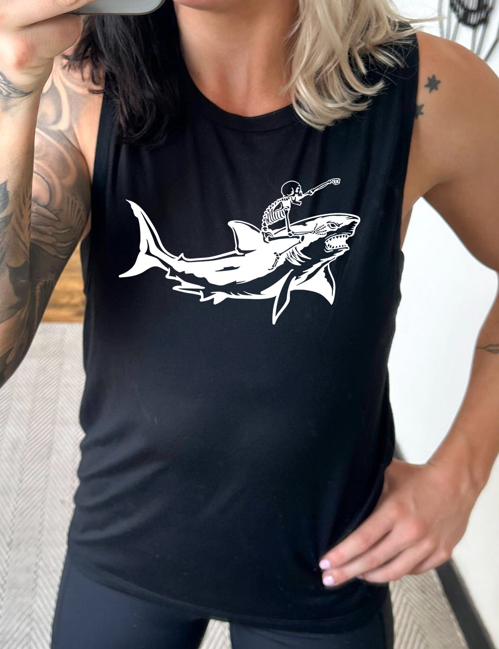 black muscle tank with the graphic of a skeleton, with its fist pointing onwards and its mouth open, riding a shark, who has a fierce expression