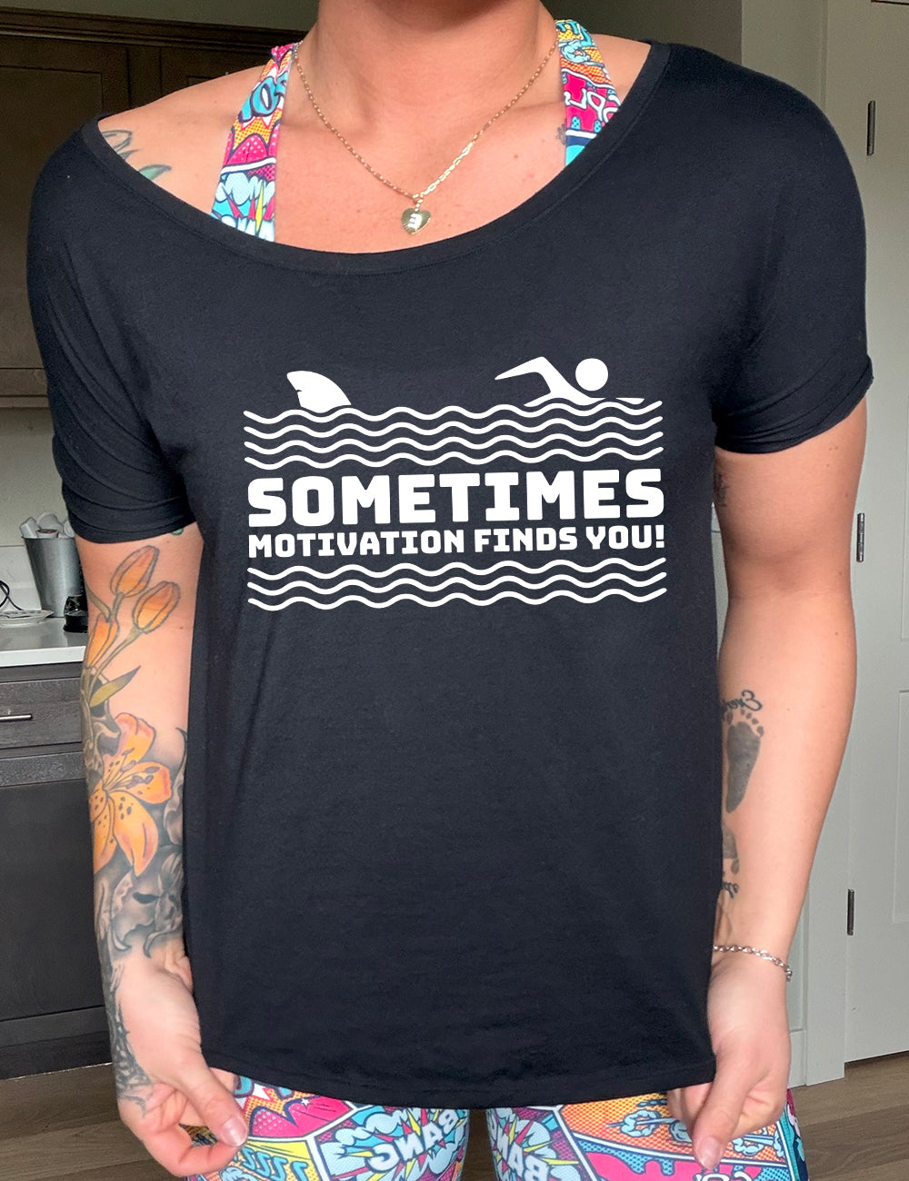 black slouchy tee with the words "sometimes motivation finds you!" in between the lined graphic of waves of the sea, and on top of the waves is the graphic of a figure swimming away from a fin in the water behind