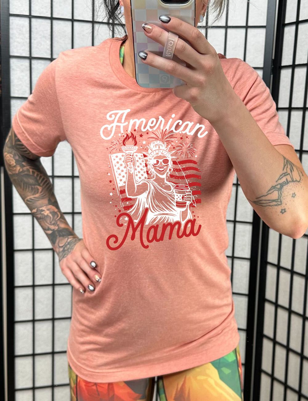 sunset unisex tee with the graphic of the statue of liberty in front of an American flag, fireworks above, wearing American flag sunglasses, holding a American flag coffee cup, with the words "American Mama" surrounding it