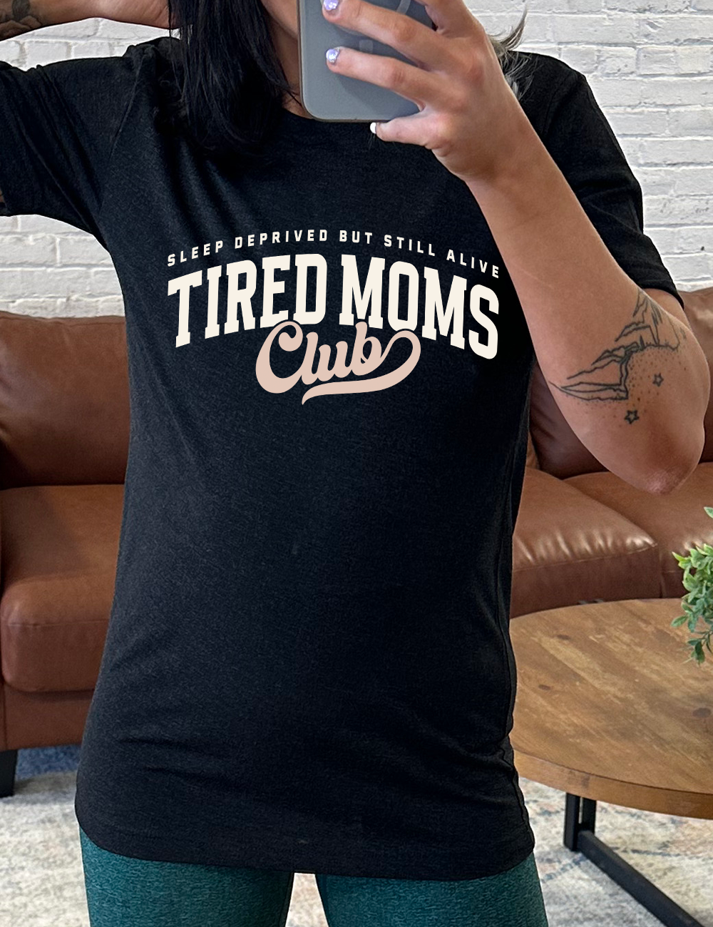 black unisex tee with the words "sleep deprived but still alive" in smaller letters, and "tired moms club" in large letters