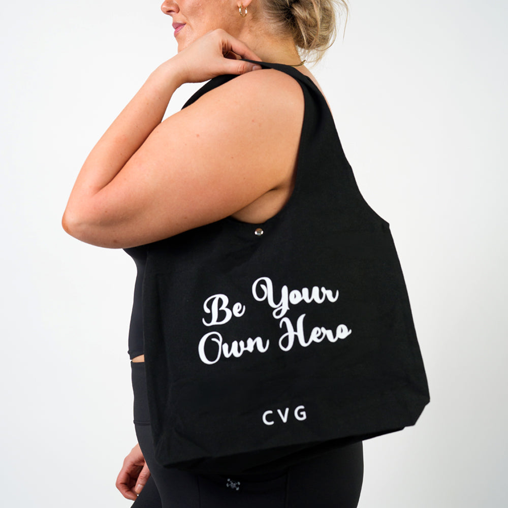 model holding a black tote bag with the saying "be your own hero"