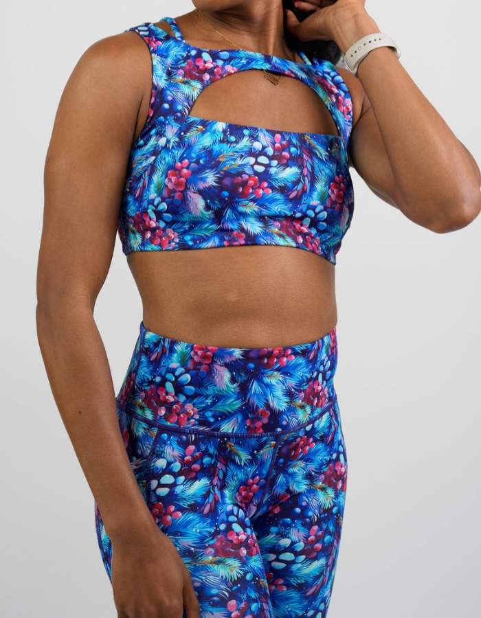 front of the blue sports bra with pine cones, holly berries, and pine branches against a white background