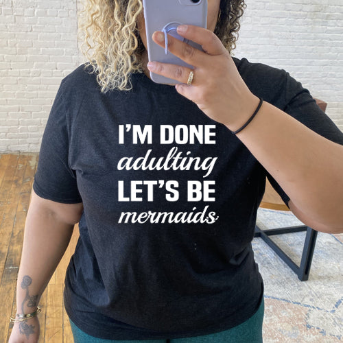 Model wearing a black unisex tee with the saying "I'm done adulting lets be mermaids" in white