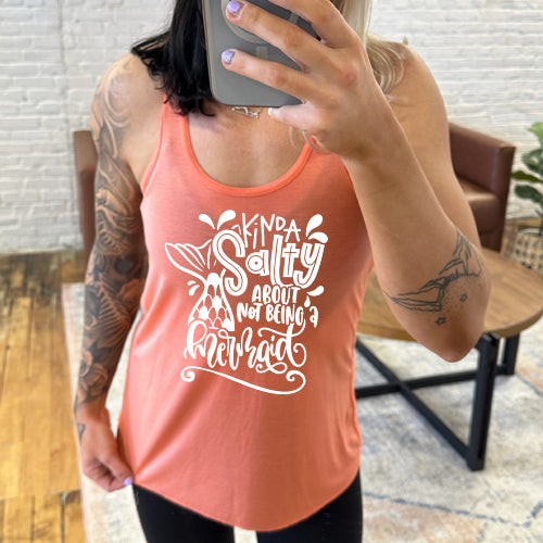 Model wearing a coral racerback tank with the saying "kinda salty about not being a mermaid" in the center