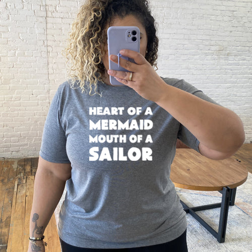 Model wearing a grey unisex tee with the saying "heart of a mermaid mouth of a sailor" in white