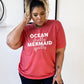 Model wearing red unisex tee with the saying "ocean shells mermaid spells" in white in the center