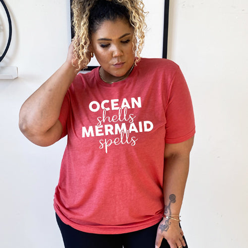 Model wearing red unisex tee with the saying "ocean shells mermaid spells" in white in the center