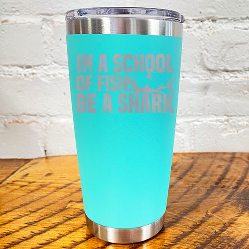 20oz teal blue tumbler with silver saying "in a school of fish be a shark" with a shark cartoon over the word "fish"