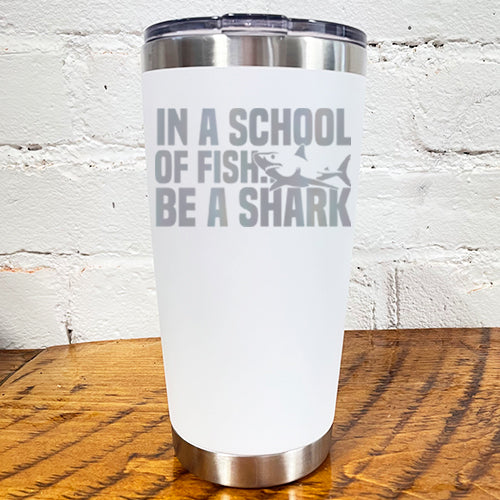 20oz white tumbler with silver saying "in a school of fish be a shark" with a shark cartoon over the word "fish"